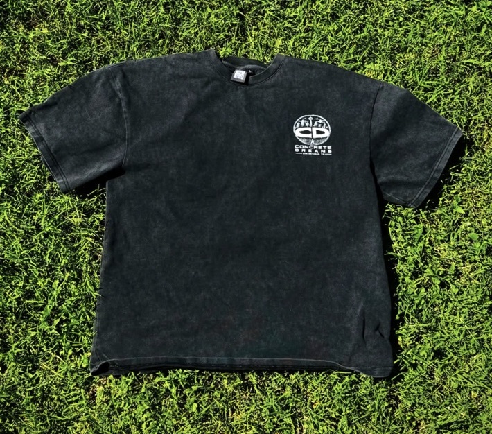 Washed black boxy short sleeve tee with a small white circular CD city logo on the left chest, laid flat on green grass